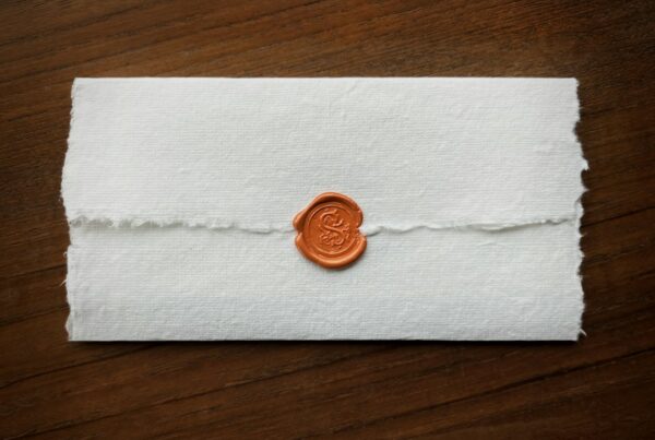 A sheet of handmade paper folded and closed with a single orange wax seal on a dark wooden desk