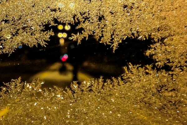 Frost crystals closing in across a dark window pane at night, leaving a small clear patch showing a street and traffic lights beyond