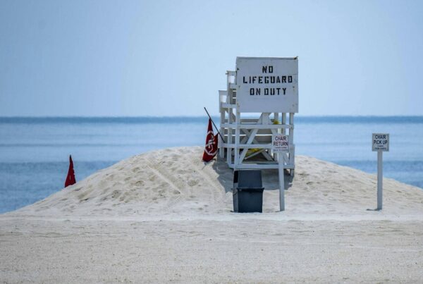 An empty white lifeguard stand on a sandy beach with a sign reading NO LIFEGUARD ON DUTY and red warning flags beside it